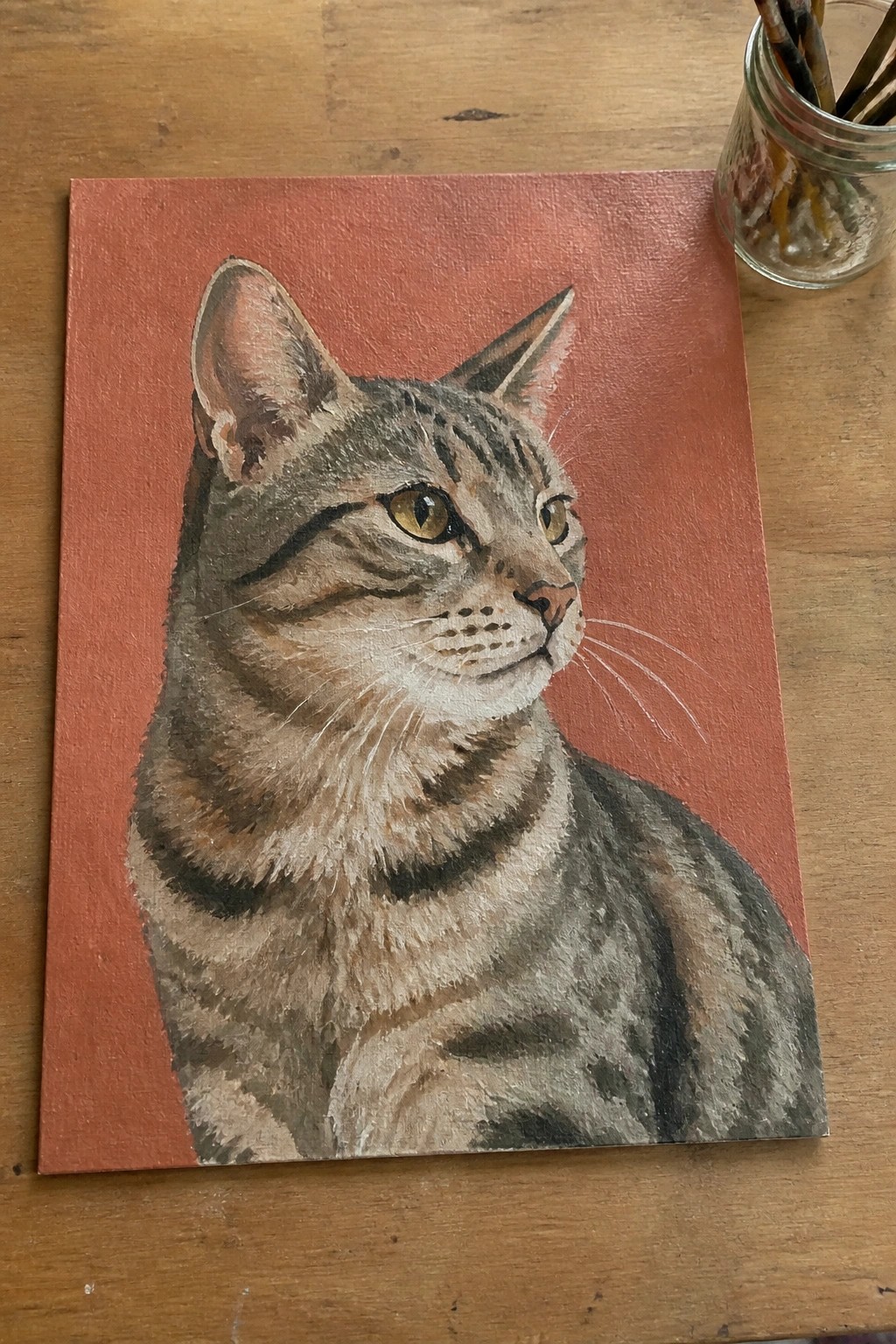 A realistic acrylic painting of a tabby cat on a solid red-orange background with a jar of brushes visible in the corner.