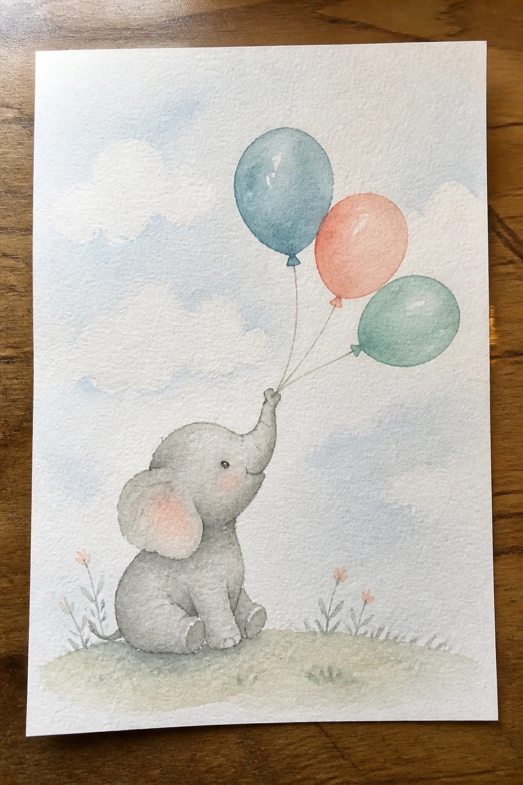 Watercolor of baby elephant sitting on grass holding blue, pink, and green balloons