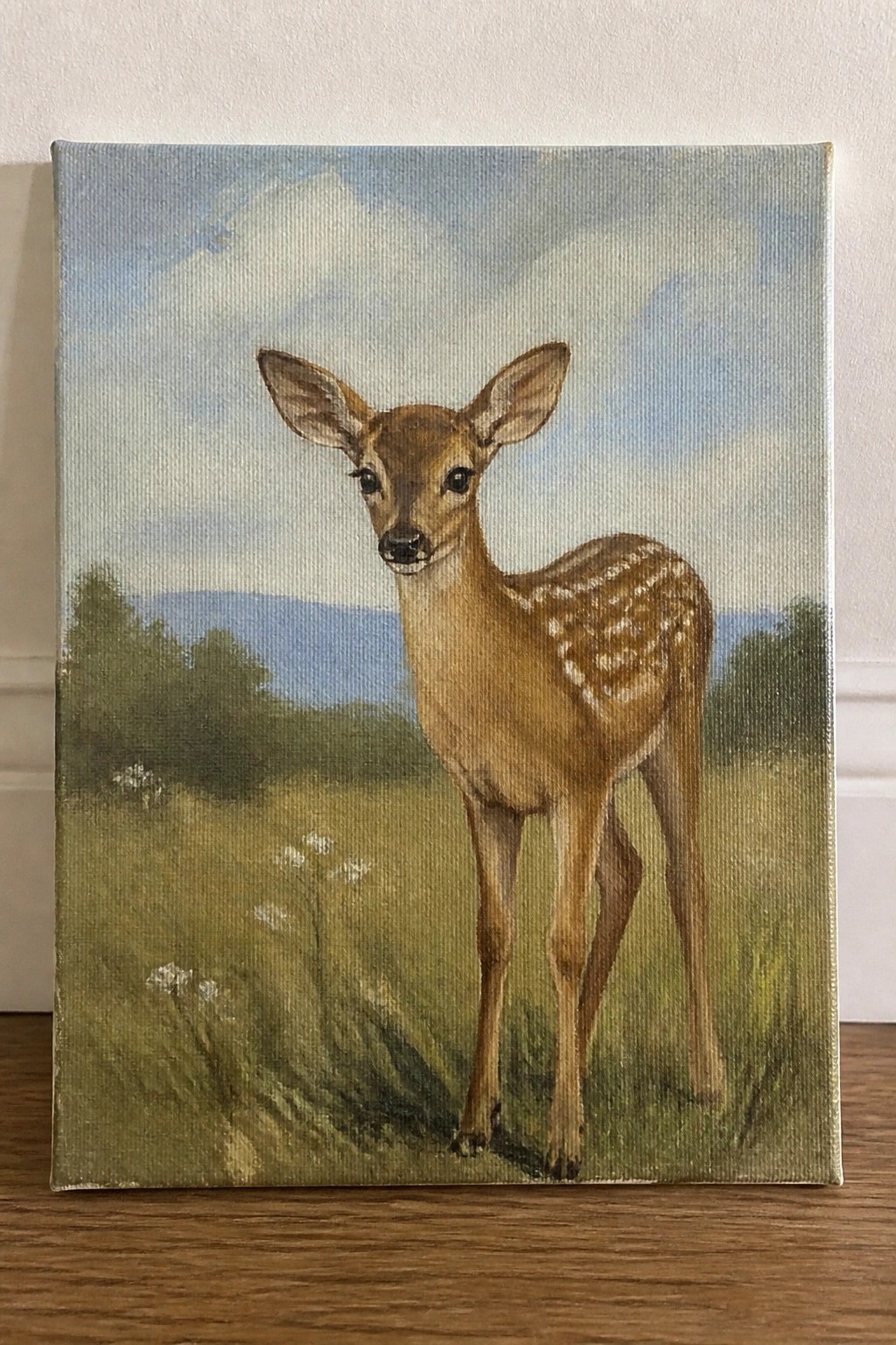 Realistic painting of a spotted fawn standing in a grassy field with mountains.