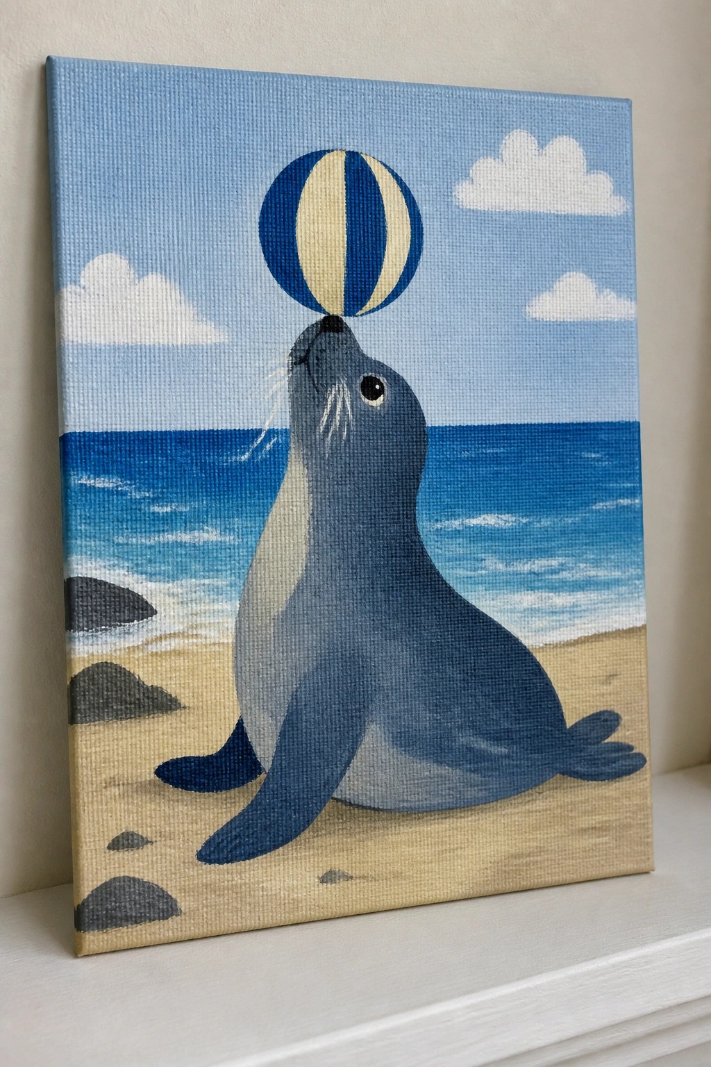 Blue-gray seal balancing striped ball on sandy beach with ocean waves.
