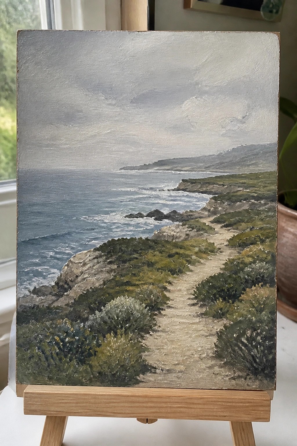 Oil painting on easel of coastal path overlooking rocky cliffs and ocean waves under gray sky