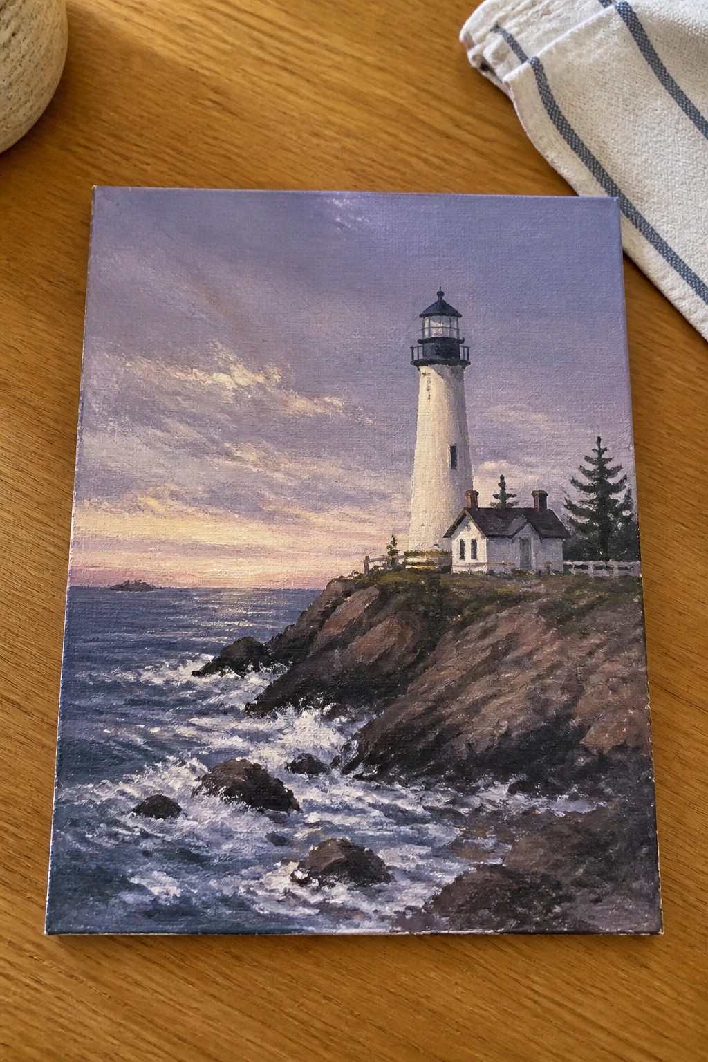 Oil painting of white lighthouse on rocky cliff by crashing waves at sunset.