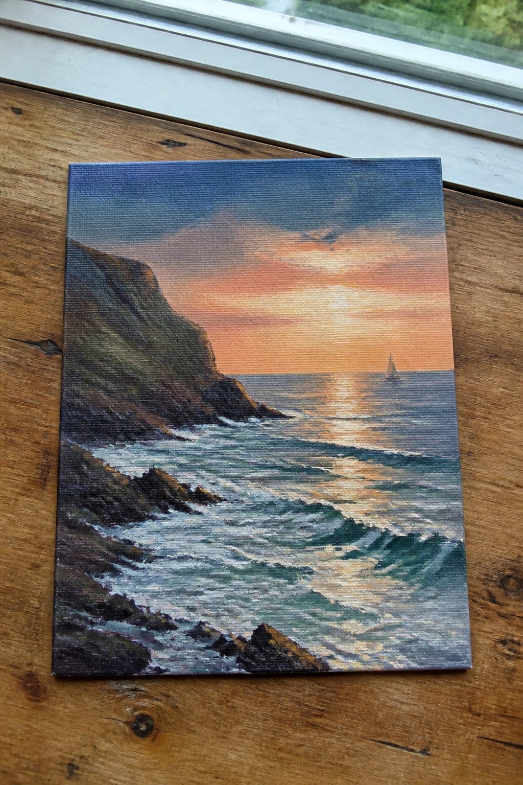 Canvas painting of sunset over ocean waves, rocky cliffs, and distant sailboat on wood.
