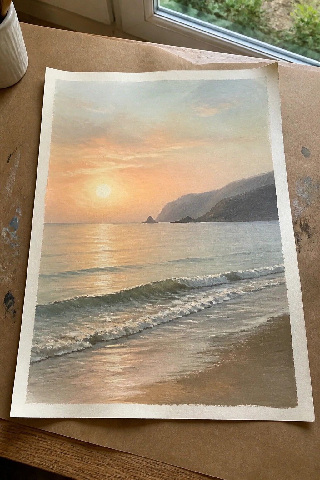 Watercolor painting of sunset over ocean with waves on sandy beach and distant cliffs on table