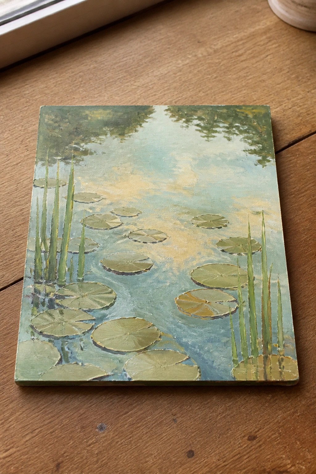 Impressionistic painting of green lily pads and reeds on a misty pond, on wood.