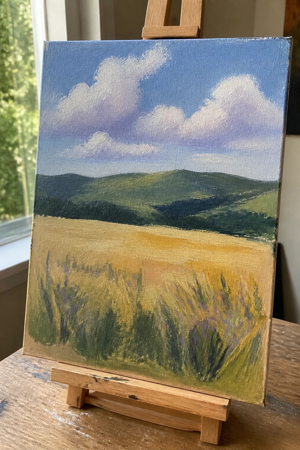 Landscape painting on wooden easel shows rolling green hills and golden field under blue sky.