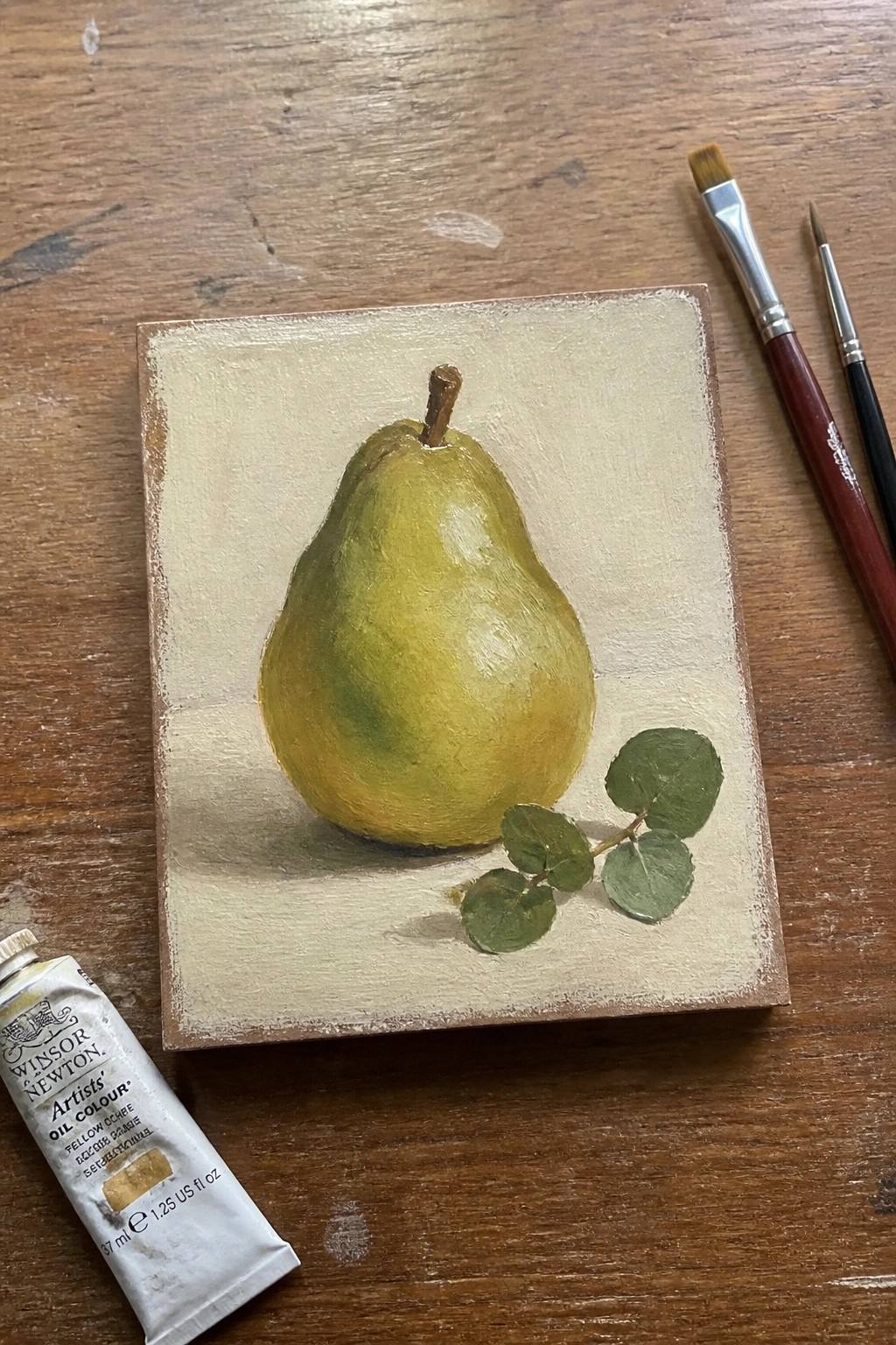 A small oil painting of a green-yellow pear with stem and a few green leaves on a beige background, next to paintbrushes and a tube of yellow ochre paint on a wooden table.