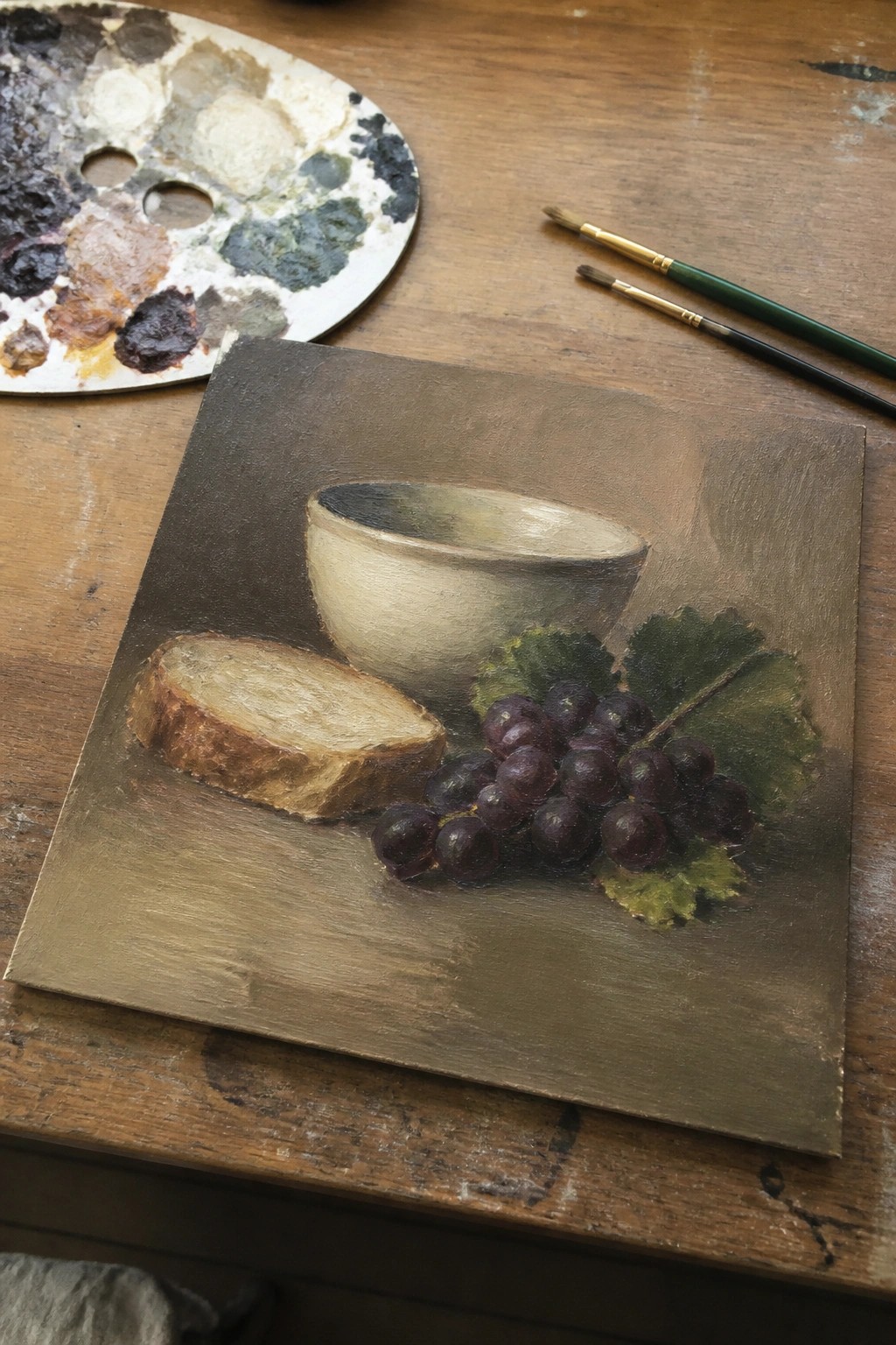 Still life painting of bread, grapes, and bowl beside paint palette and brushes