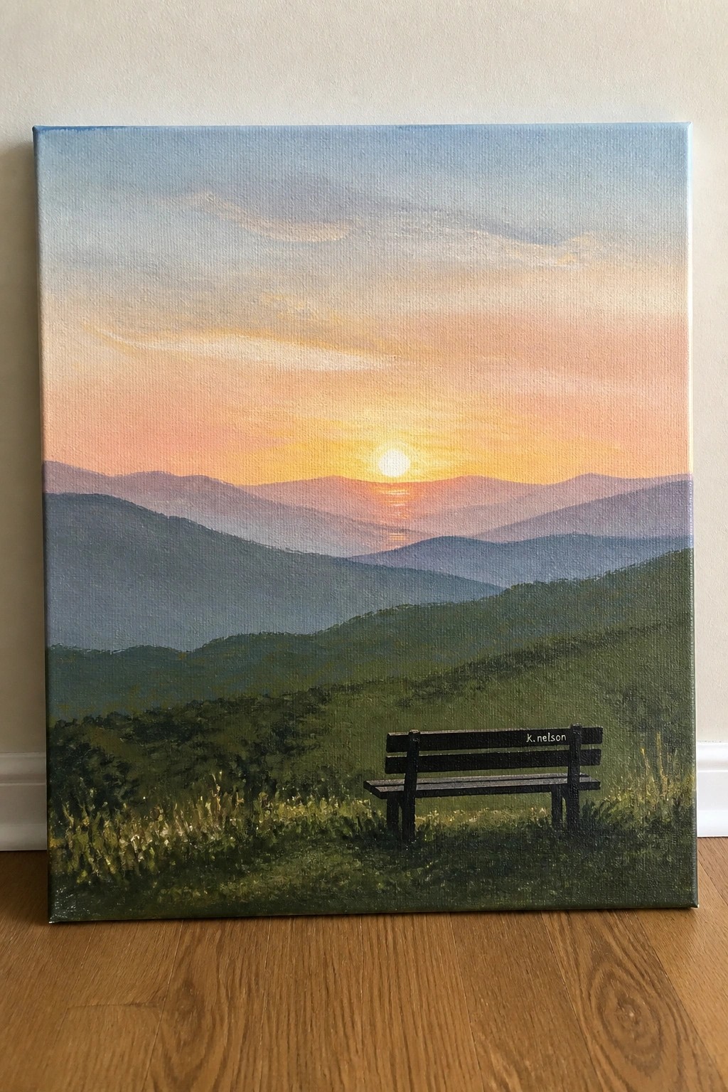 Sunset painting of layered mountains with wooden bench on grassy hill.