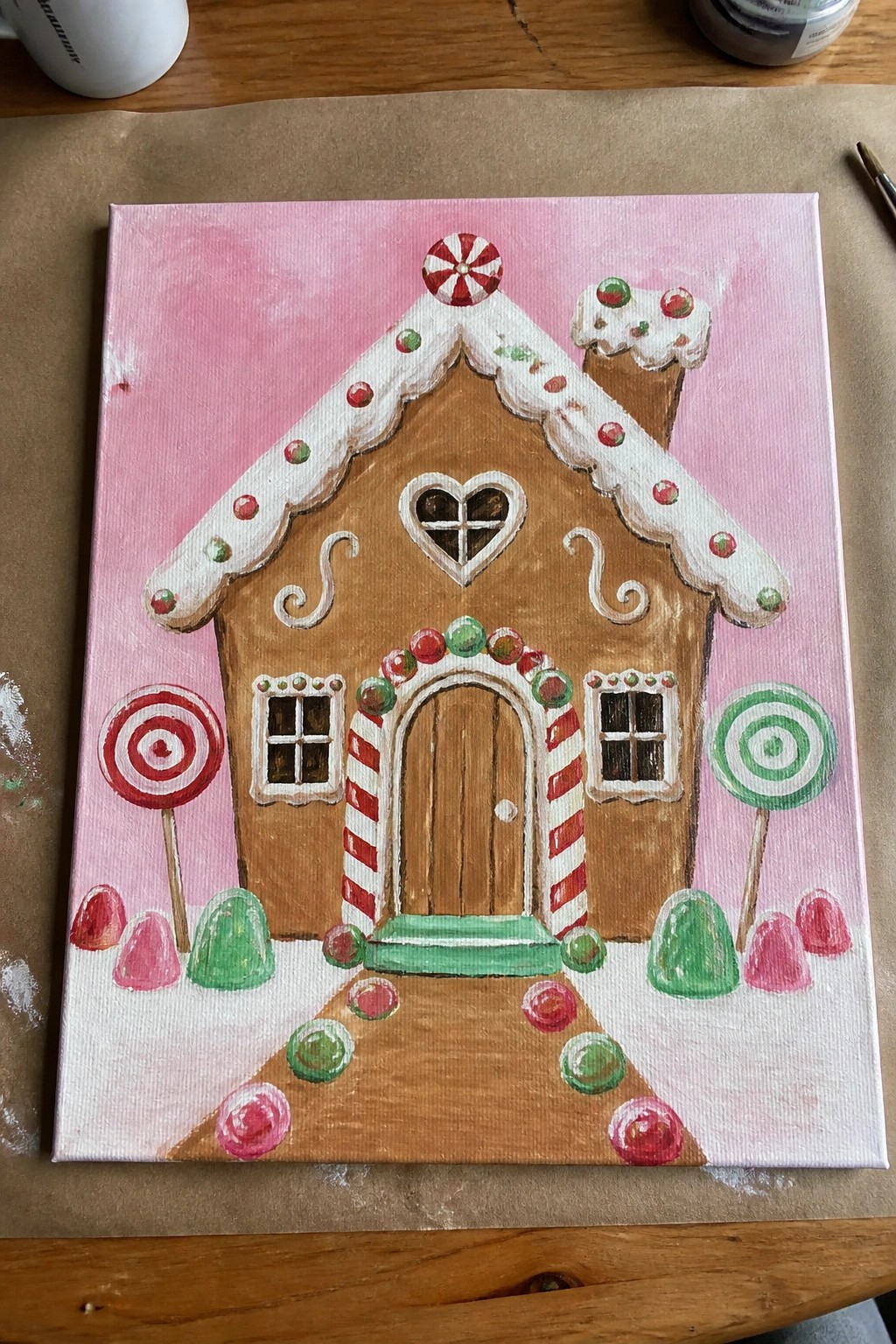 Festive gingerbread house painting with candy decorations, heart window, and lollipops on pink canvas.