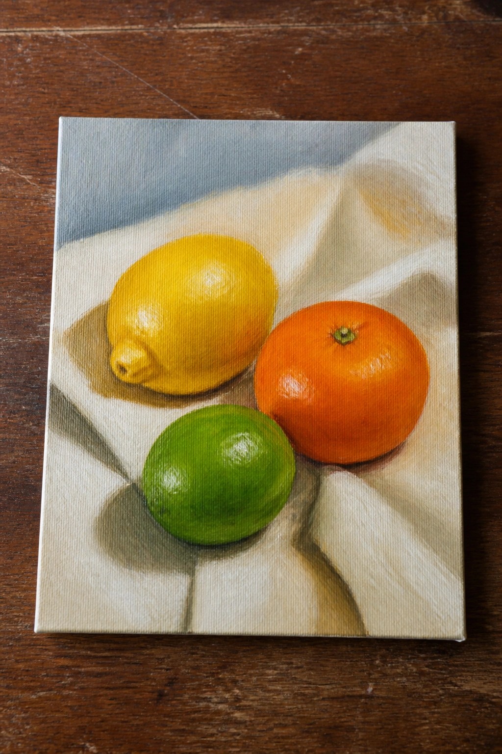 Still life painting of yellow lemon, green lime, and orange on white draped cloth