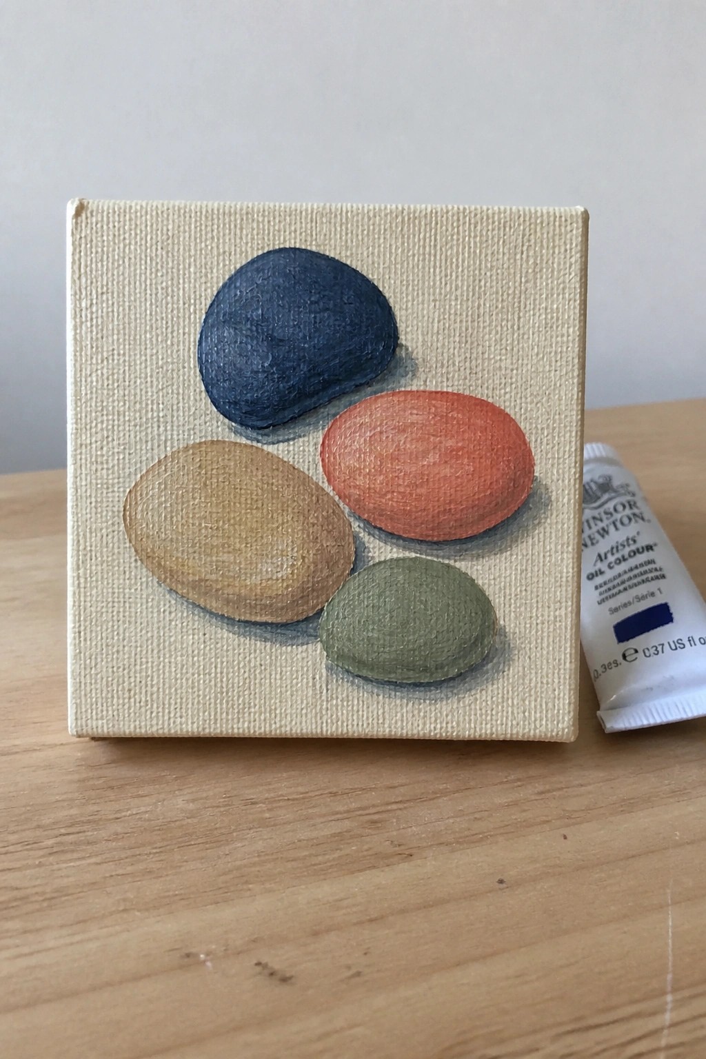 Small canvas shows four painted pebbles in blue, orange, tan, and green beside paint tube