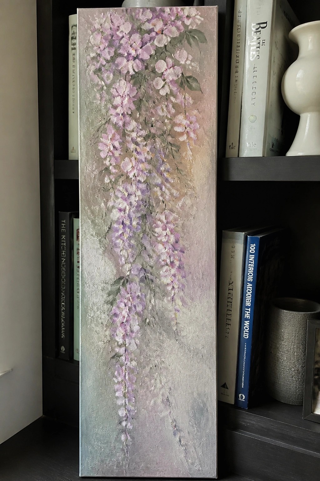 Tall painting of cascading purple wisteria flowers on bookshelf with books
