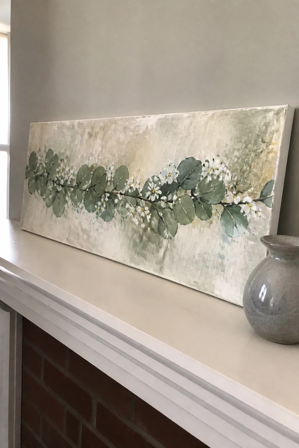 Floral painting with green leaves and white blooms on white mantel beside vase