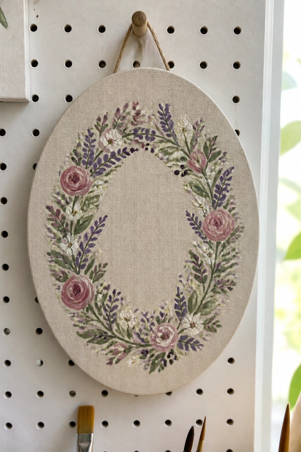 Oval canvas floral wreath with pink roses and purple blooms hanging on pegboard