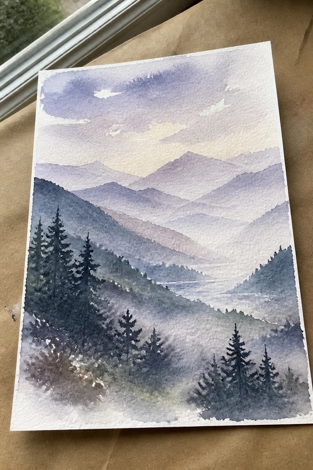 Watercolor painting of misty layered mountains in purple hues with dark pine trees foreground.