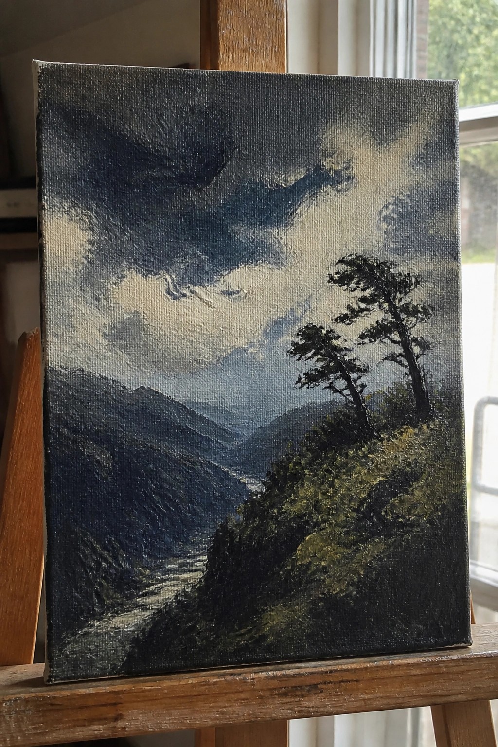 Landscape painting of dark mountains, river, and trees on wooden easel
