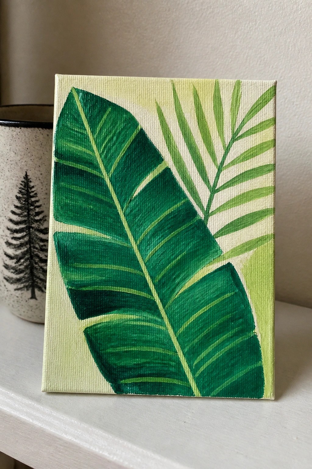 Small canvas with vibrant green tropical leaves on pale yellow beside tree mug
