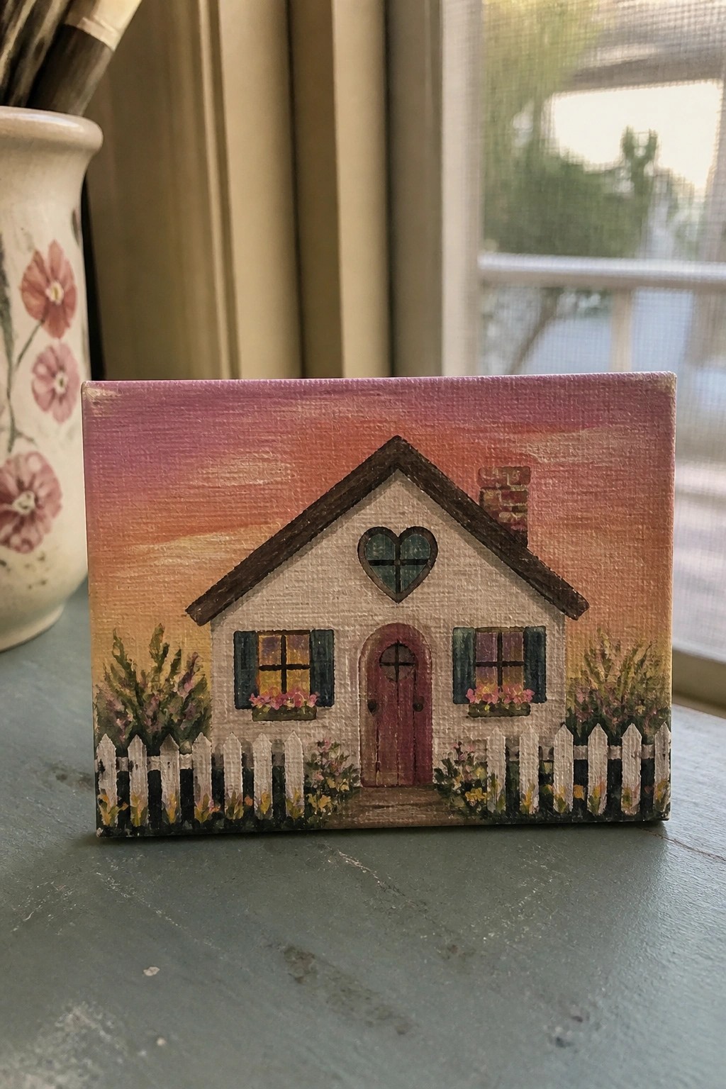 Small canvas painting of cottage with heart window, picket fence, and pink sunset sky