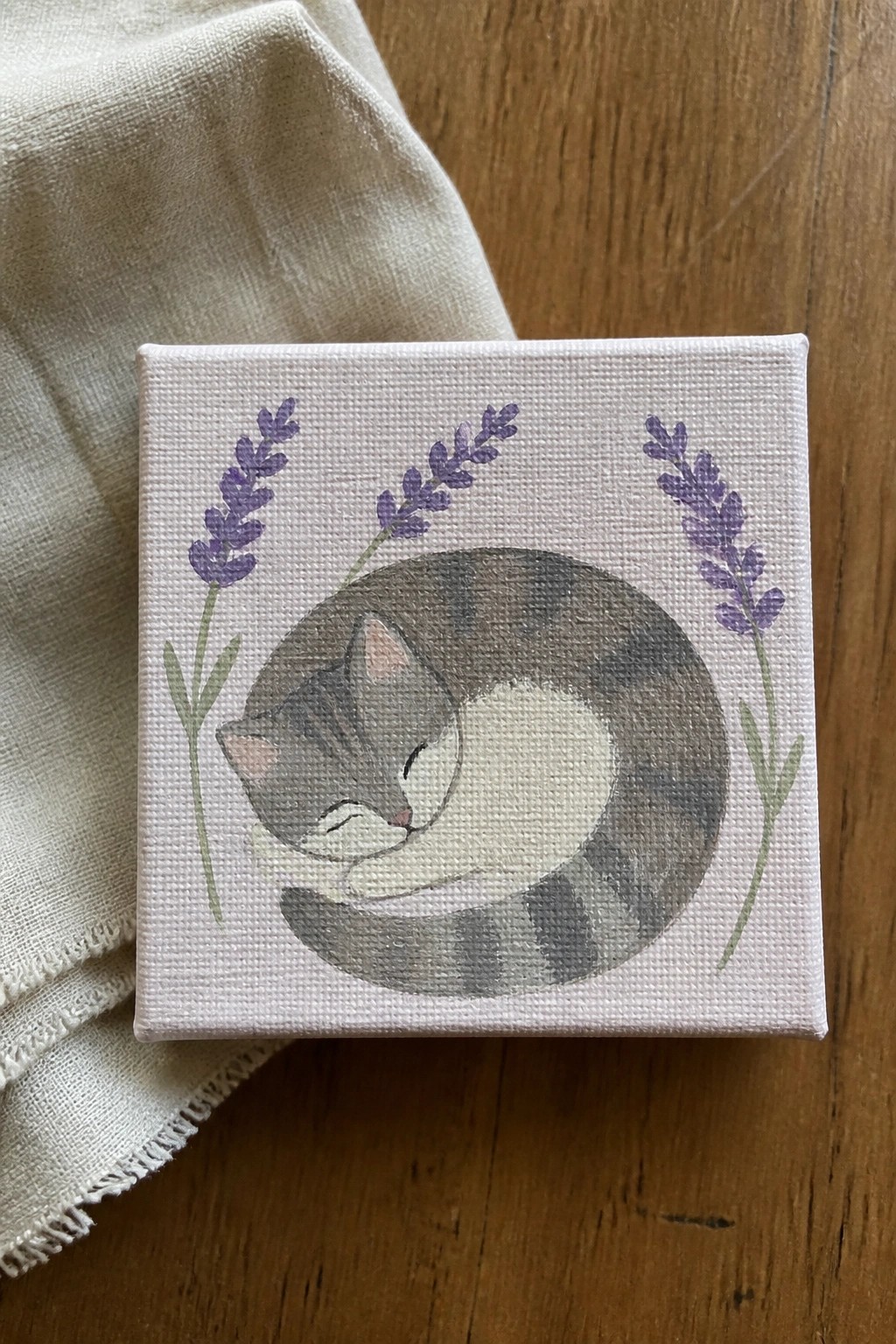 Small canvas painting of sleeping gray tabby cat with lavender sprigs on wood.