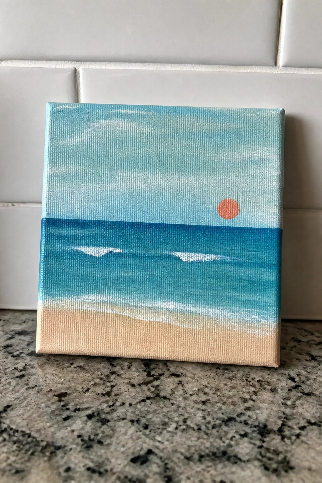 Acrylic painting on a mini canvas showing a blue gradient sky with an orange sun over a blue ocean with white waves and a sandy beach.