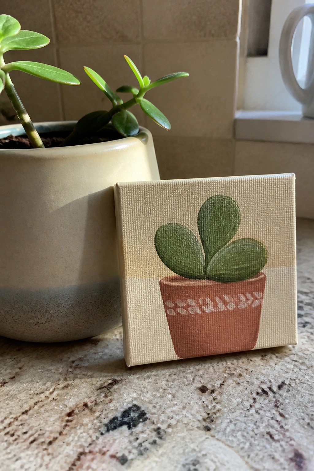 Small canvas painting of green succulent in terracotta pot beside real plant on counter.