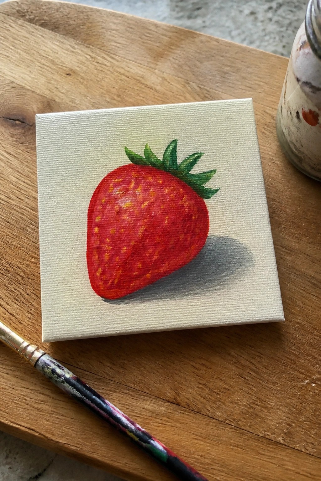 A small square canvas on wood shows a red acrylic strawberry with green leaves and a gray shadow, with a paintbrush nearby.