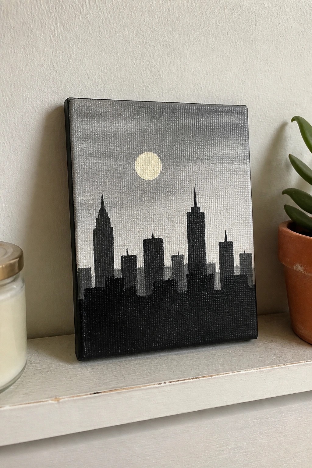 Black city skyline silhouette painting with moon on shelf beside candle and plant.