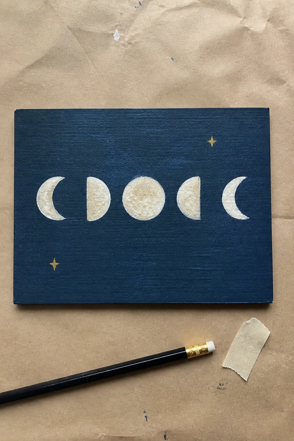Canvas painting of moon phases on dark blue with gold stars, pencil below.