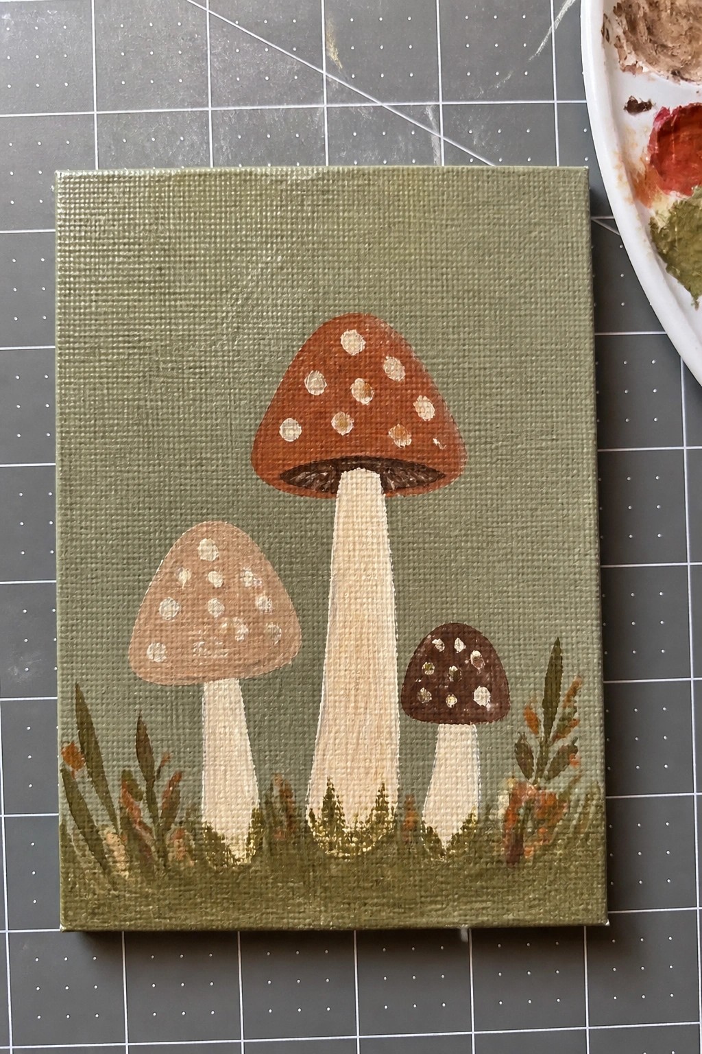 Acrylic painting of three spotted mushrooms on green canvas with foliage
