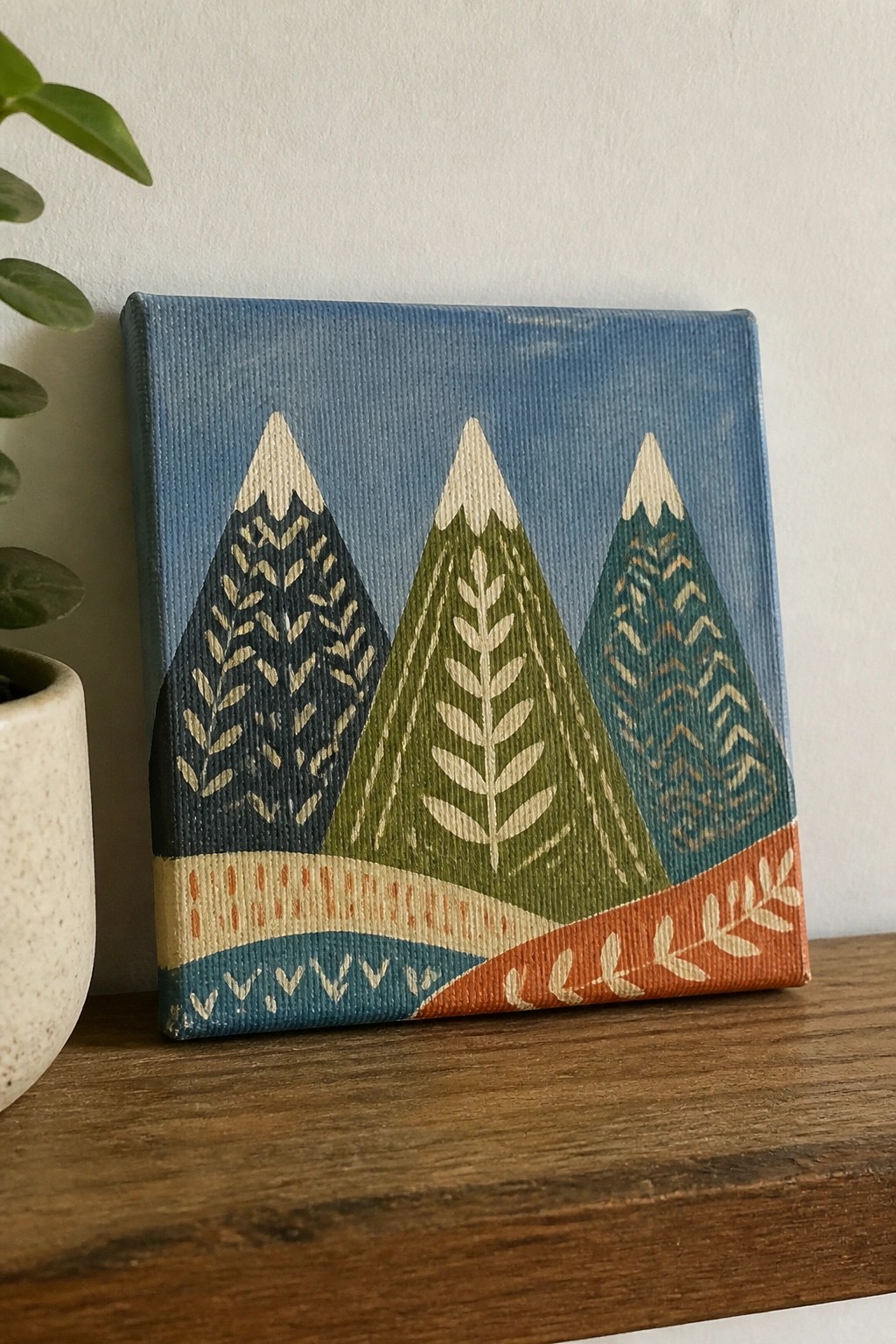 Colorful folk-art painting of three patterned mountains on wooden shelf beside plant