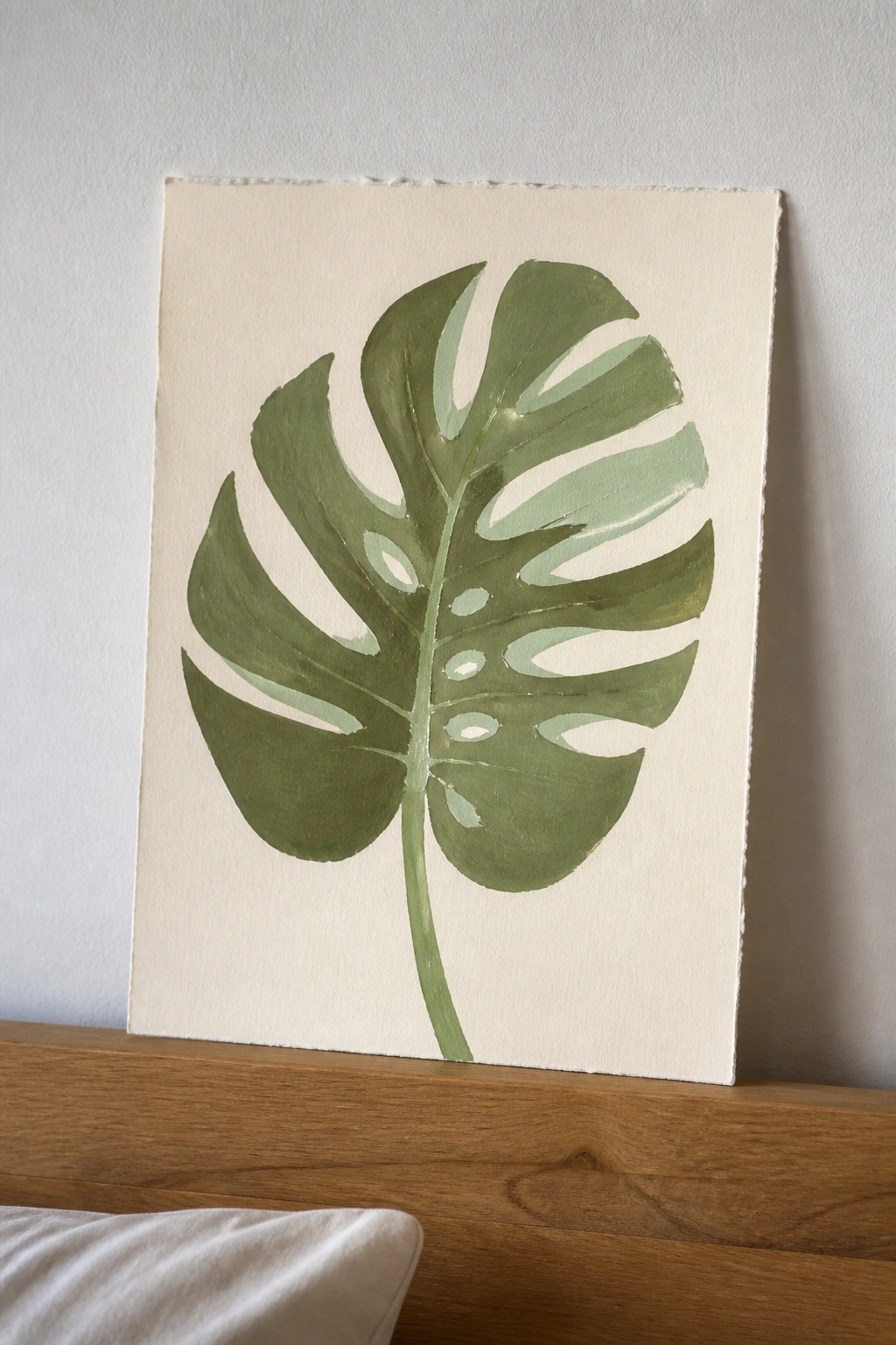 Green monstera leaf painting on off-white canvas atop wooden ledge