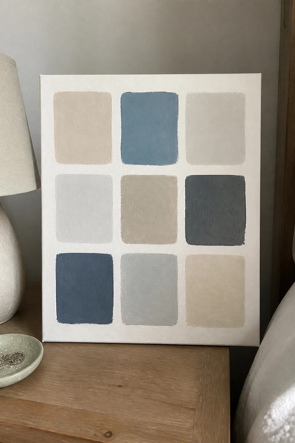 Abstract painting of nine colored squares in blue beige and gray beside white lamp on wood table