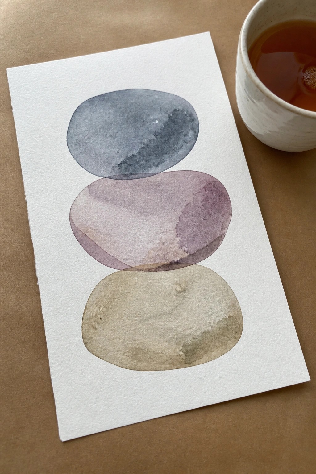 Watercolor painting of three stacked ovals beside a cup of tea on brown surface.