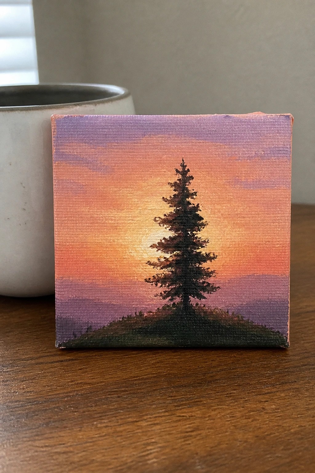 Small canvas of silhouetted pine tree against orange-purple sunset beside white mug
