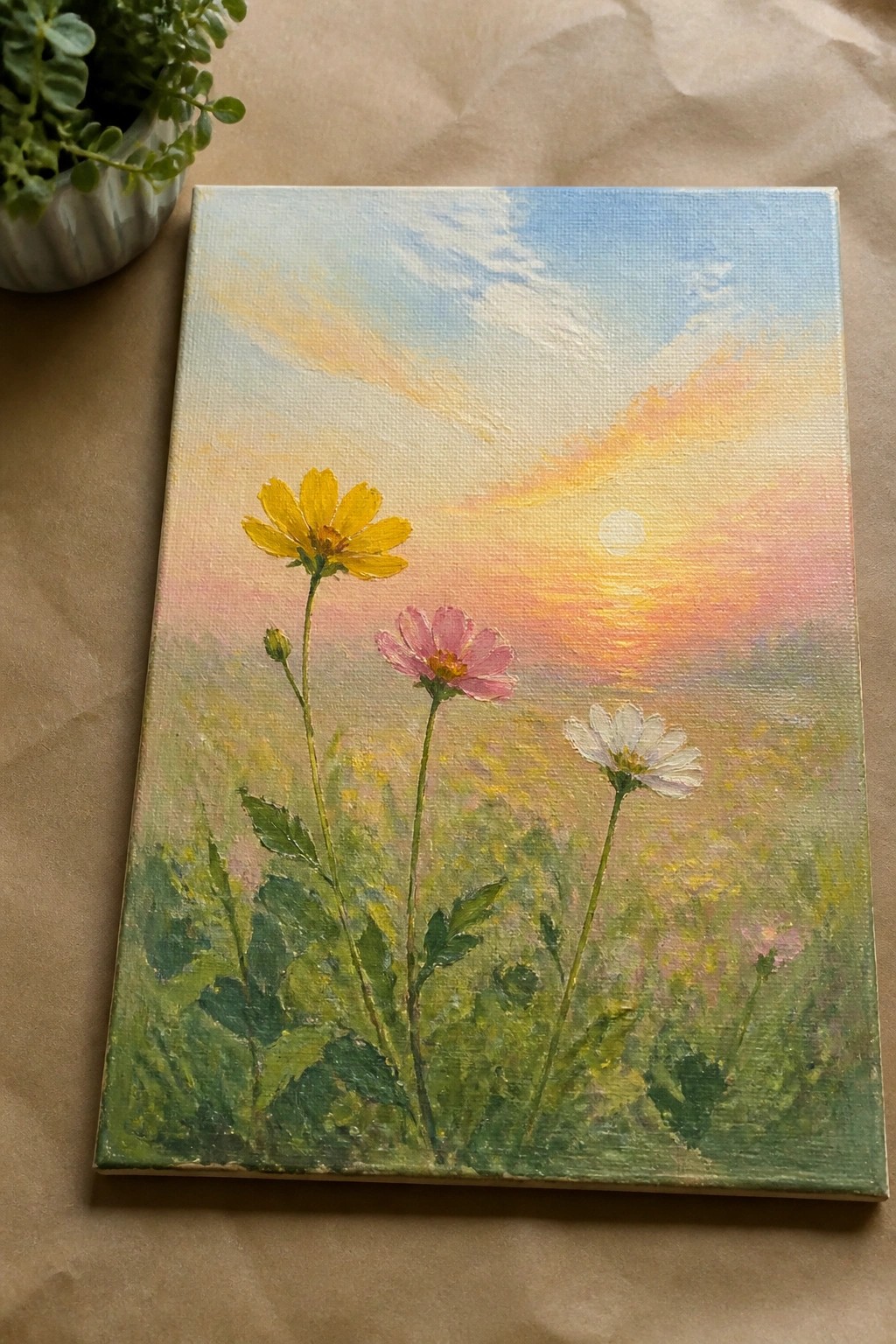 Oil painting on canvas showing a yellow daisy-like flower, a pink cosmos, and a white daisy in green grass with a glowing sunrise and sun visible in the background.