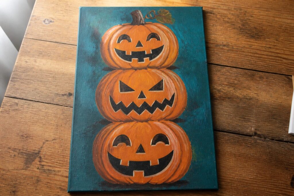 Three stacked orange jack-o'-lanterns with carved faces on teal canvas.