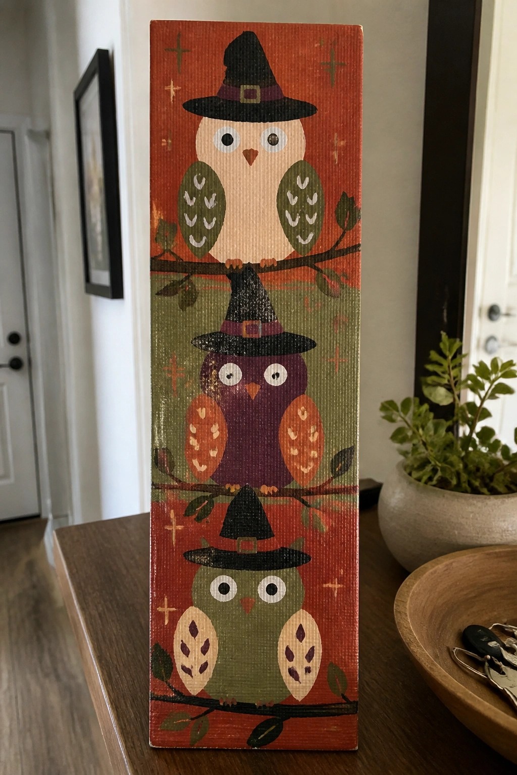 Three stacked owls in black witch hats on orange-green wooden plaque.