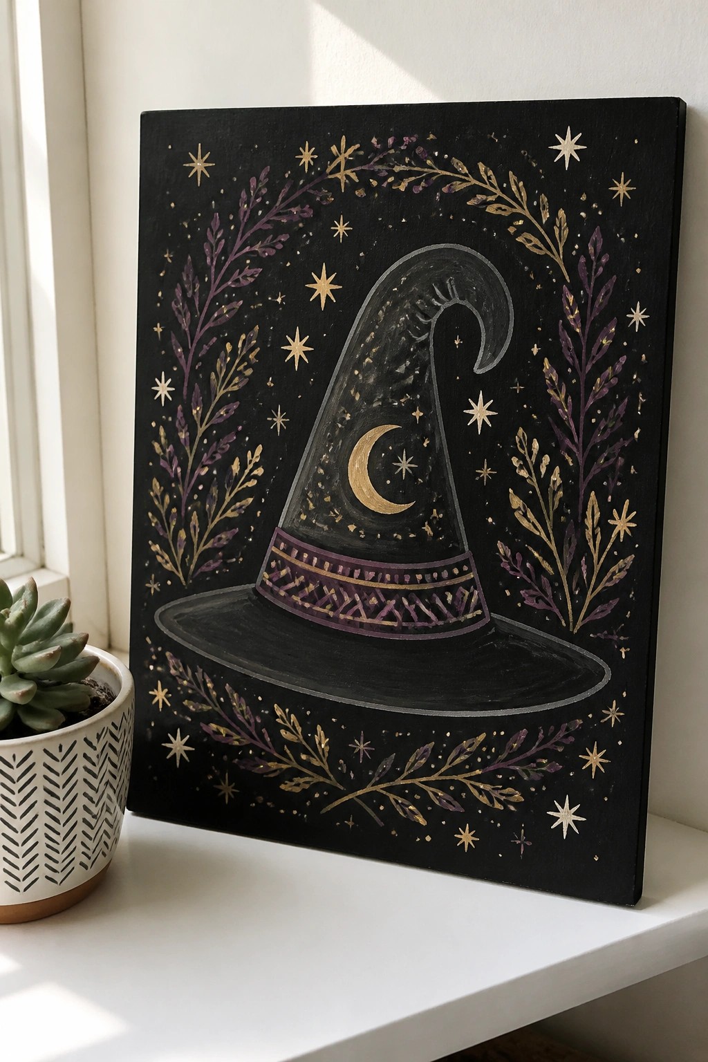 Black witch hat painting with moon, stars, and gold-purple foliage beside succulent plant.
