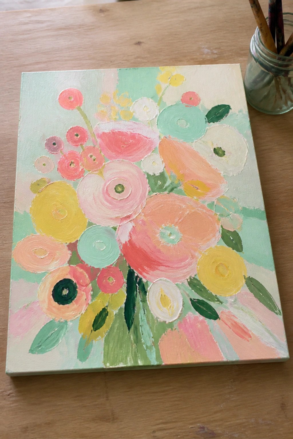 An acrylic painting of a colorful flower bouquet with pink, yellow, orange, and mint blooms on a light green and cream background sits on a wooden table next to a jar of paintbrushes.
