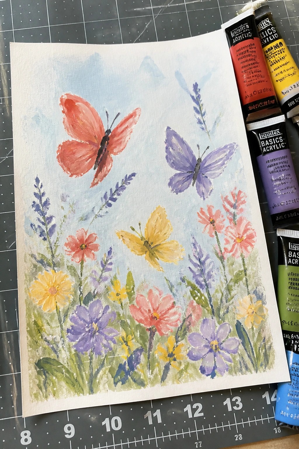 Watercolor painting of colorful butterflies amid flowers with acrylic paint tubes on mat