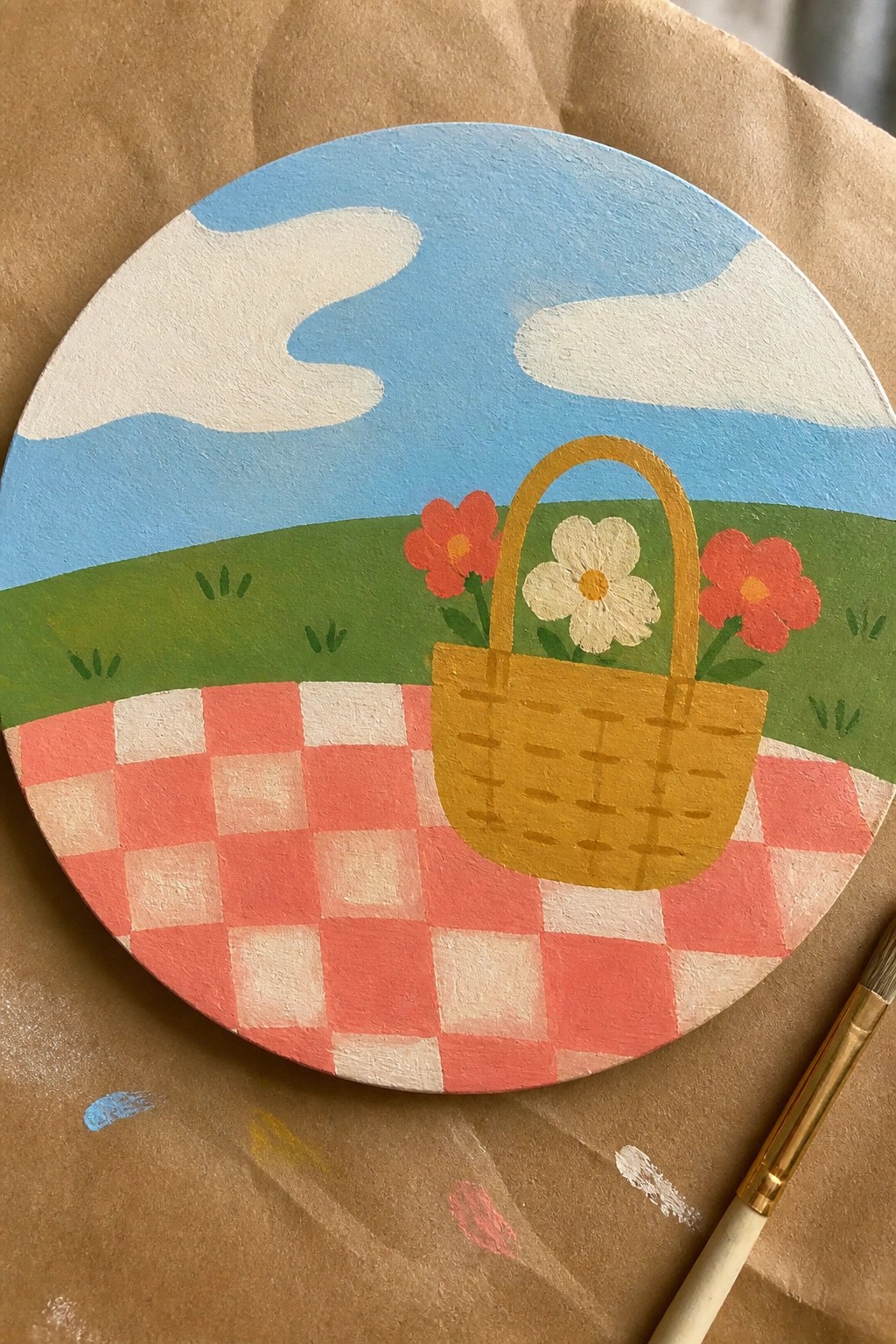 Circular painting of yellow picnic basket on red checkered blanket with flowers and blue sky