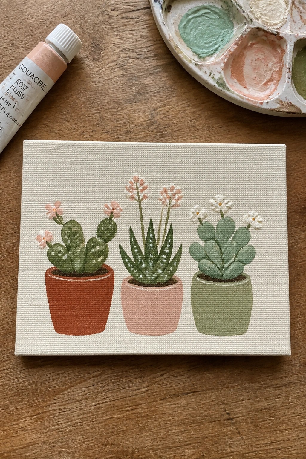 Three potted succulents with flowers on small canvas beside gouache paints