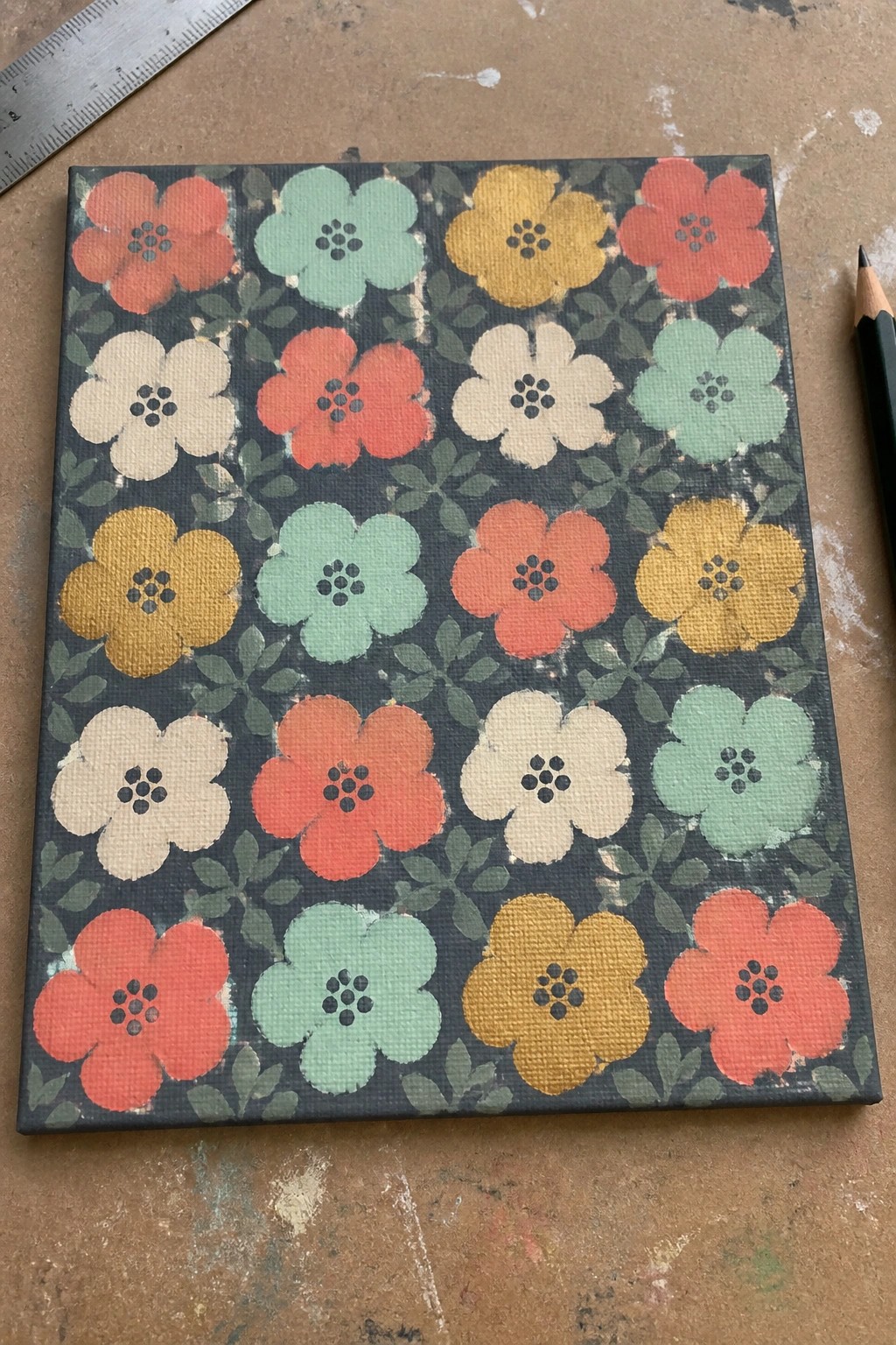 Square canvas with retro floral pattern in red, yellow, mint, and cream beside ruler and pencil.