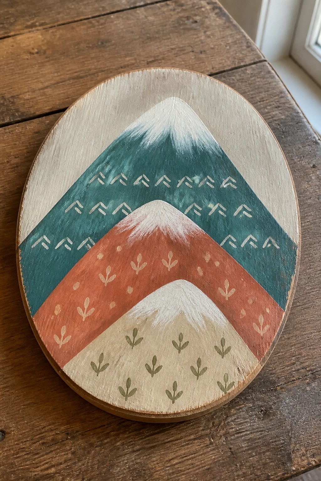 Round wooden plaque with layered teal, red, and beige mountains and decorative patterns.