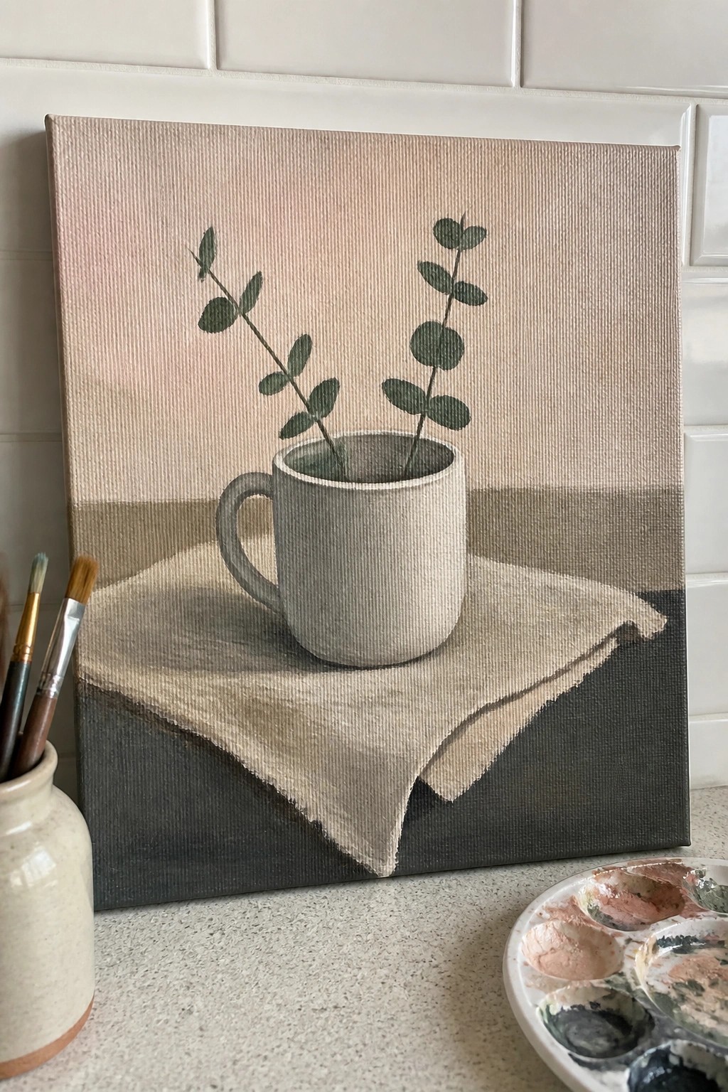 Canvas still life of gray mug with eucalyptus on beige cloth beside palette