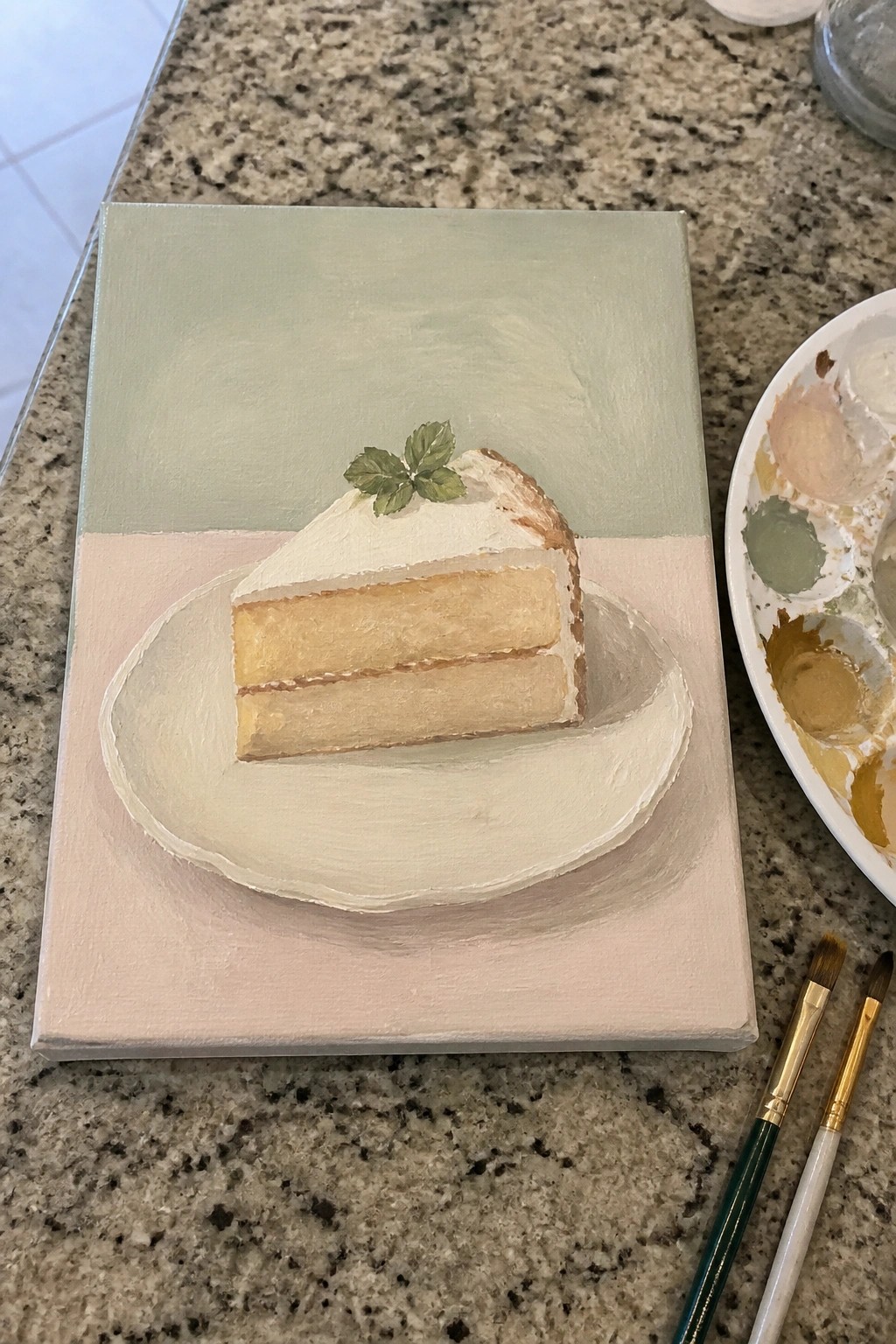 Painting of cake slice on plate beside palette and brushes on granite counter.