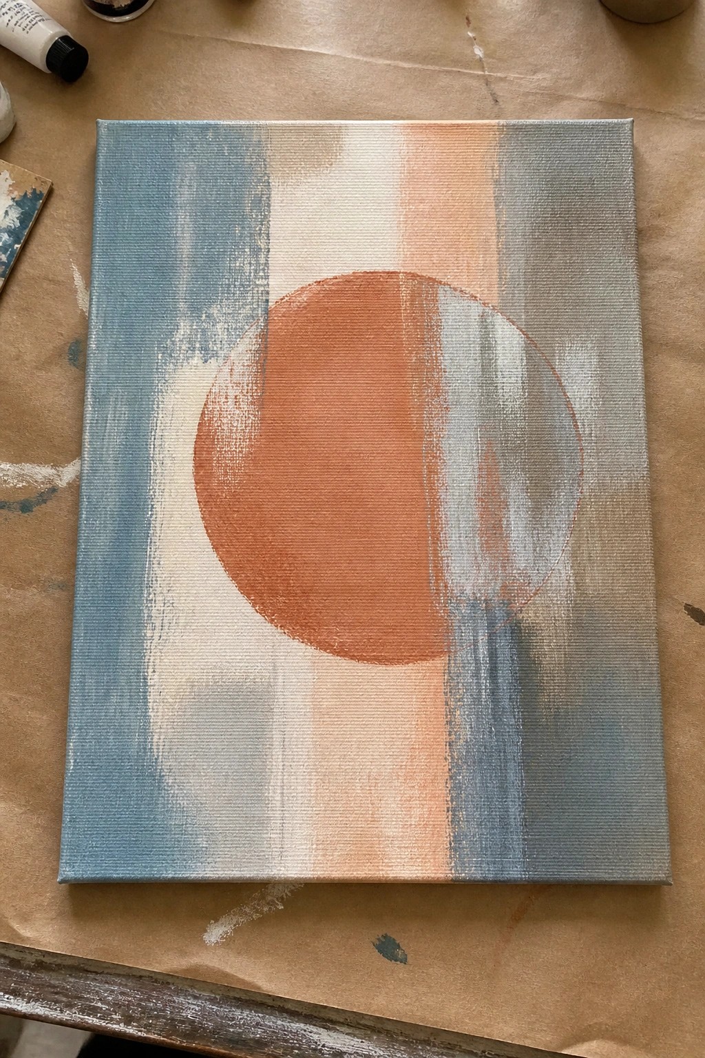 Abstract canvas with orange circle over blue, white, peach, and gray vertical stripes on brown paper