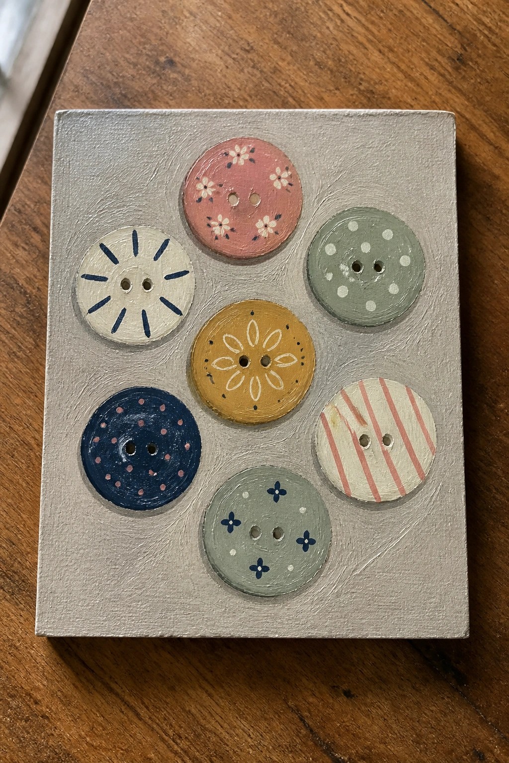 Seven colorful patterned buttons painted on a gray square canvas.