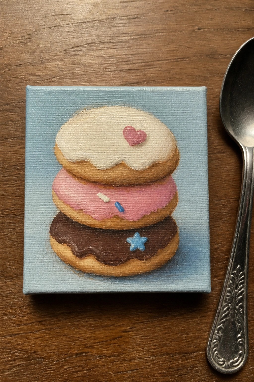 Mini canvas painting of three stacked donuts with icing and sprinkles beside spoon